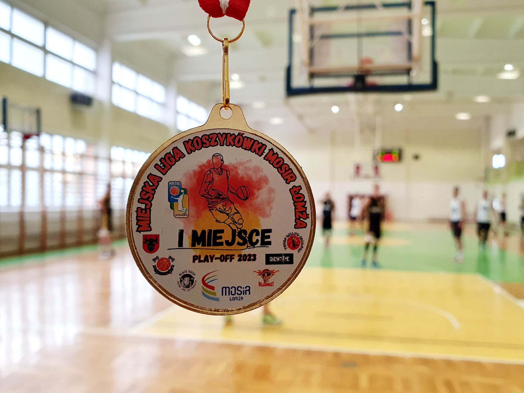 zdjęcie przedstawiające medal, a w tle zawodnicy koszykówki w sali sportowej